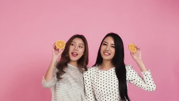 幸福两个迷人的女人在粉红色的背景下跳舞。亚洲美女拿着切片橙色水果健康cear概念