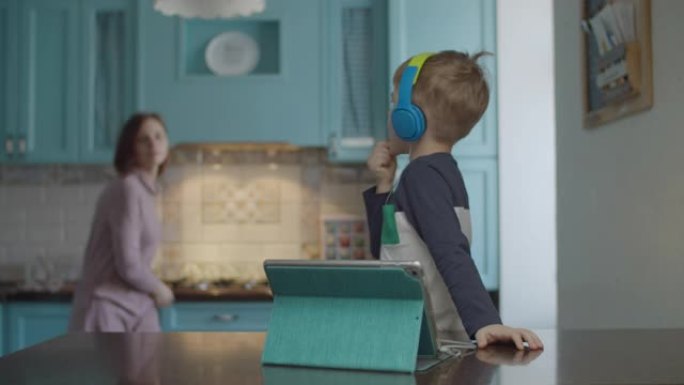 穿着彩色耳机的金发学龄前男孩在厨房的桌子上使用平板电脑。母亲来寻求帮助