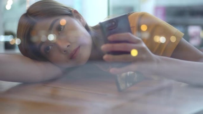 散焦的年轻美丽孤独的亚洲女人坐在窗边，躺在桌子上，晚上在咖啡馆使用智能手机。沮丧的少女用悲伤的眼睛看
