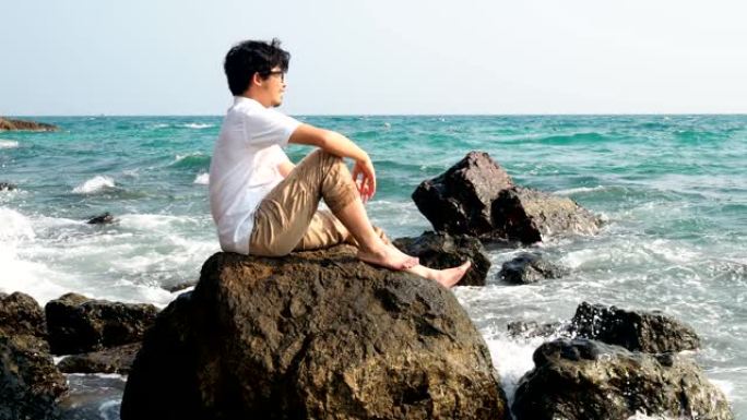 年轻轻松的亚洲男子坐在海边的岩石上