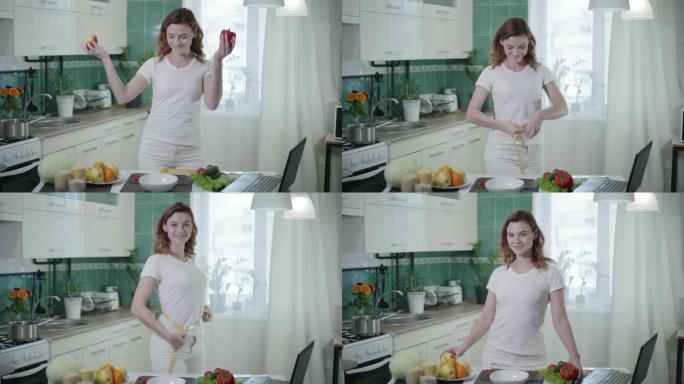 微笑的女孩在厨房里用健康食物在相机上摆姿势后快乐，用卷尺测量她的腰部