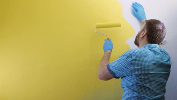 疲倦的人用黄色的左手用滚筒画白色的墙壁。他穿着格子蓝色衬衫和手套。慢动作和中等镜头