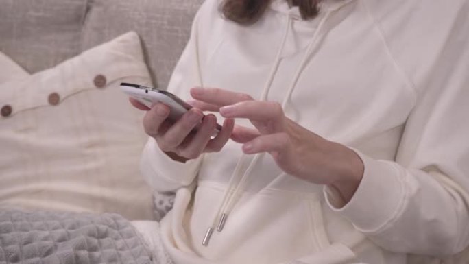 放松概念: 女人坐在沙发上，用智能手机从前面拿走一条毯子，同时手指在屏幕上滑动