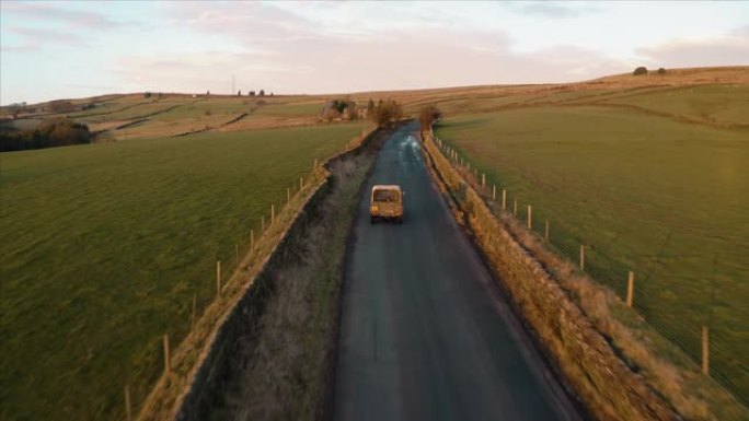 在美丽的日落时分，一辆车在后面有一只狗的车辆穿越英国乡村的农地时，航拍镜头