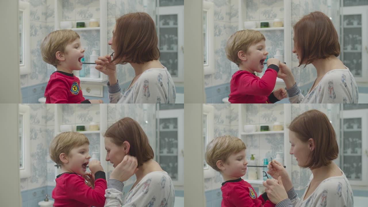 年轻的母亲帮助穿着红色睡衣的学龄前男孩用牙线刷牙。有趣的家庭沐浴程序。母子俩在浴室里大笑。