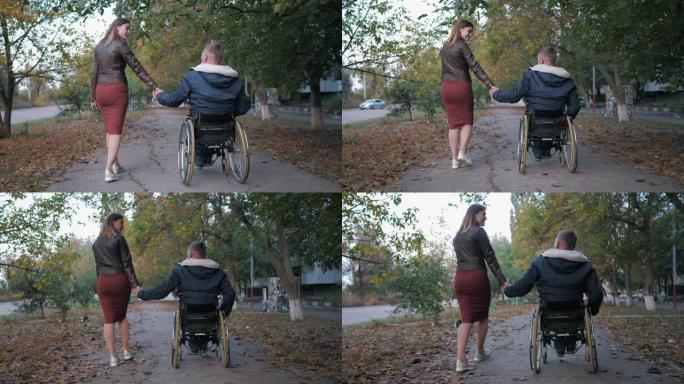 敬爱的坐在轮椅上的残疾人在树木中的秋天公园散步时，握着妻子的手