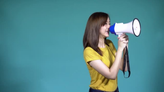 亚洲快乐美丽可爱的年轻女子青少年兴奋她使用扩音器新闻公告看着工作室的相机，孤立在蓝色背景，4k视频溶
