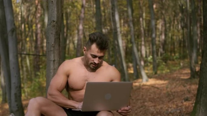 强壮的裸体躯干家伙坐在秋天的公园里，手里拿着笔记本电脑。