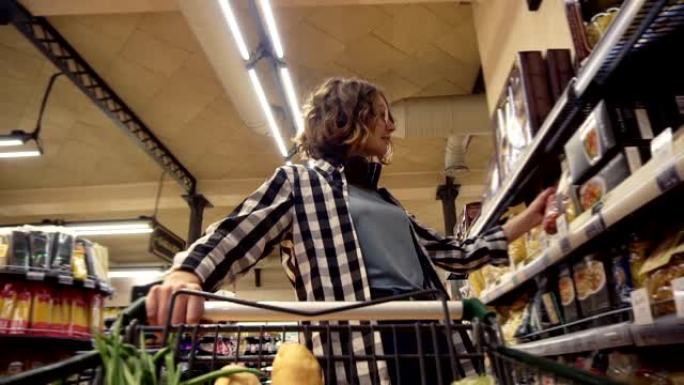 穿着格子衬衫和脖子上戴着耳机的微笑女人正走在杂货店里，转向购物车，里面有食物。站在分类的前架子上，拿