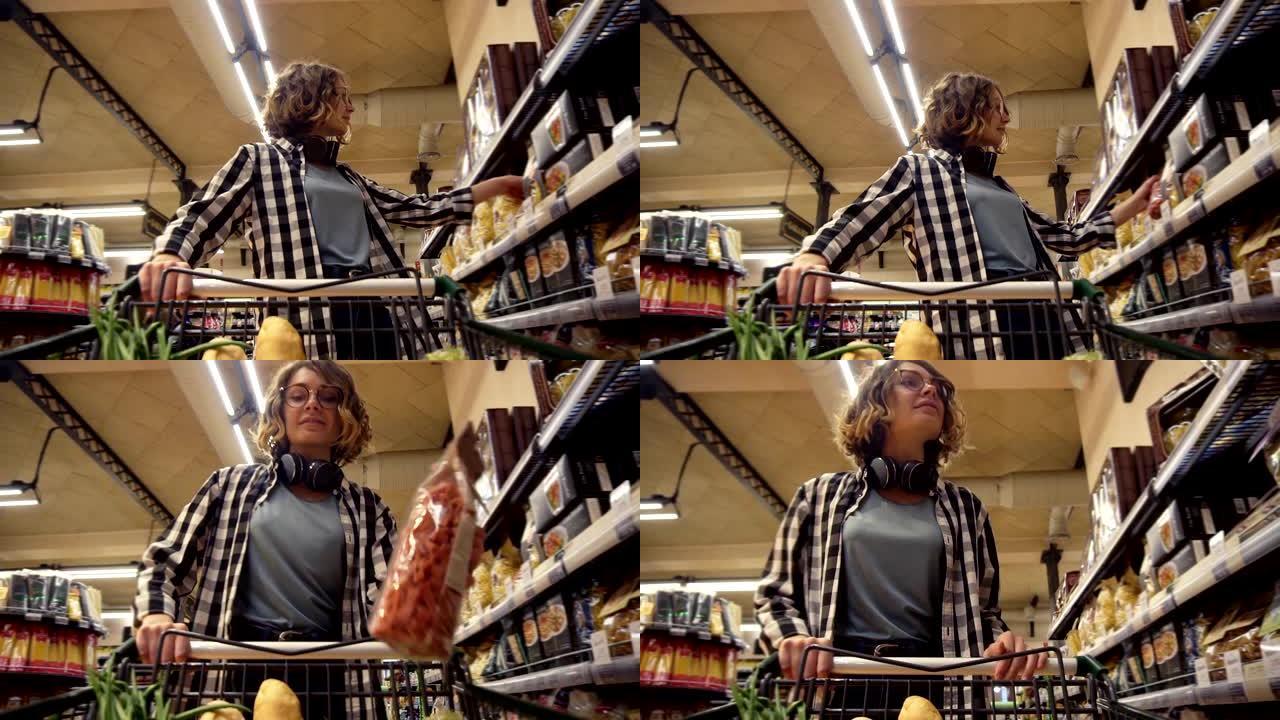 穿着格子衬衫和脖子上戴着耳机的微笑女人正走在杂货店里，转向购物车，里面有食物。站在分类的前架子上，拿