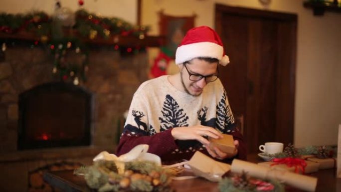 戴着圣诞老人帽子的快乐男人打开壁炉附近的圣诞礼品盒，花环闪烁。微笑的家伙收到了一个带礼物的包裹，享受