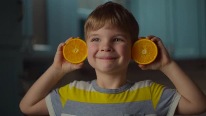 学龄前男孩手里拿着两半橙色水果，把它放在耳朵上，微笑着看着相机。橙色水果婴儿耳朵在慢动作。