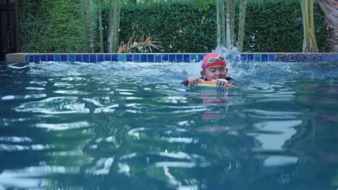 穿着游泳衣的快乐亚洲男孩穿游泳镜在室内游泳池度假村使用浮板。节日快乐。慢动作