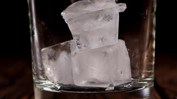 在黑暗的木制背景上，冰块落入空玻璃杯中的特写镜头。