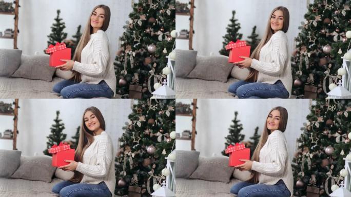 迷人的年轻女性微笑着拿着红色礼品盒靠近圣诞树。在红色相机上拍摄