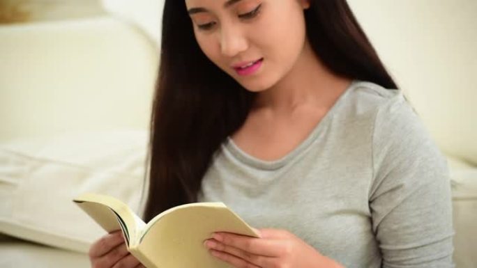 亚洲女人坐在舒适的沙发上看书。放松的女人假期时间早上在温暖舒适的家中看书。