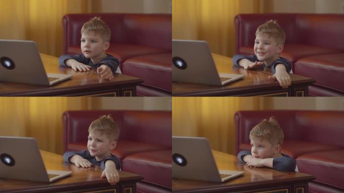 金发碧眼的学龄前男孩在笔记本电脑上打字，微笑着看着电脑屏幕。孩子坐在桌旁在家在线学习。
