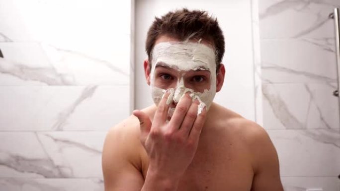 年轻英俊男子的肖像正在脸上涂上新鲜的白色厚面膜和擦洗。天然有机护肤化妆品。男人淋浴后，面部皮肤护理