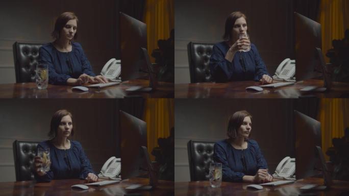 在夜间办公室工作的年轻女商人。女人在键盘上打字，用柠檬喝水。带监视器的办公桌上的工作女性。前视图。