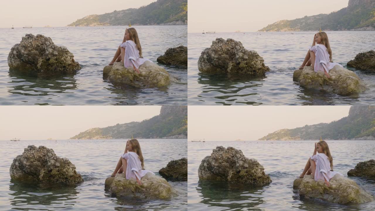 穿着白色外衣的放松女孩在水中的石头上享受海洋景观。暑假时，快乐的年轻女孩坐在阳光明媚的海滩上的海水岩