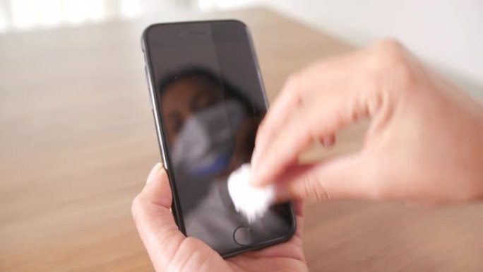 一位戴着面罩的女士正在用棉纸和酒精清洁手机，担心新型冠状病毒肺炎和冠状病毒，脸在屏幕上反射。