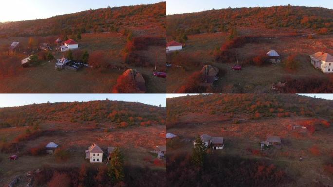 在阳光明媚的秋天，在散布着房屋和橙色落叶松森林的小山村上空前进。山间蜿蜒的山路鸟瞰图。