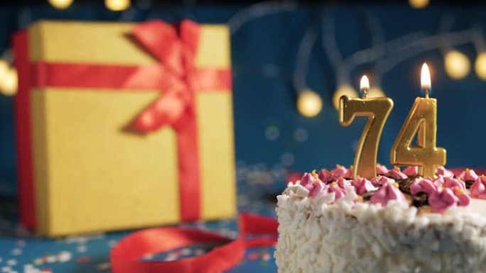 白色生日蛋糕74号点火器燃烧的金色蜡烛，蓝色背景带灯和用红丝带捆绑的礼物黄色盒子。特写