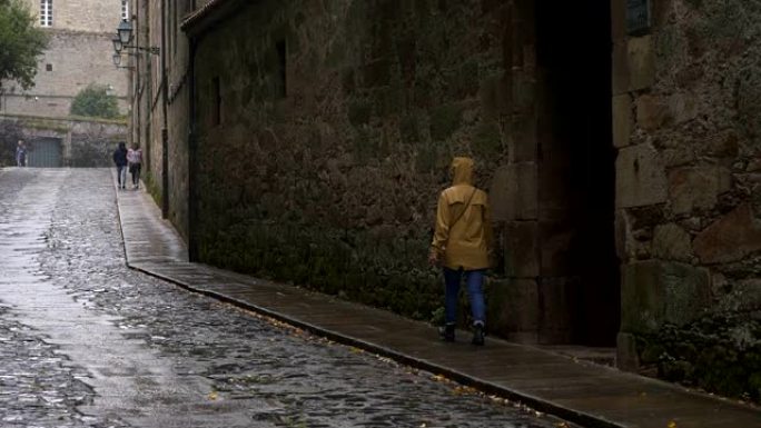 西班牙圣地亚哥德孔波斯特拉市，女旅行者穿着黄色夹克走在下雨天的街道上