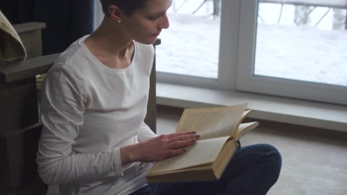 在一个寒冷的冬日傍晚，一个短发的年轻女孩坐在窗边的地板上看书。享受文学的女人。女性教育和学习。小屋的