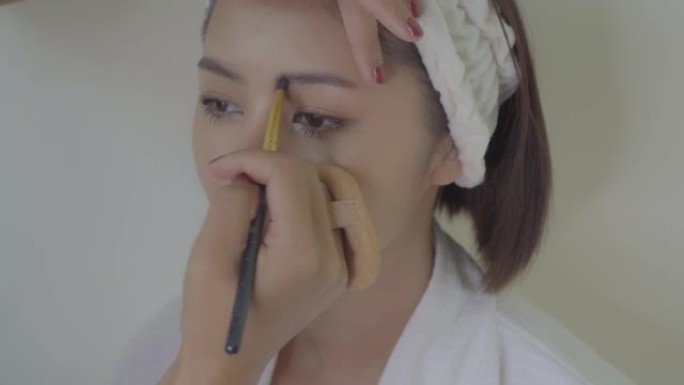 化妆师用眉刷在美女年轻的亚洲女人去派对前填充眉毛颜色。年轻的亚洲妇女留在美容院，赤裸裸地化妆