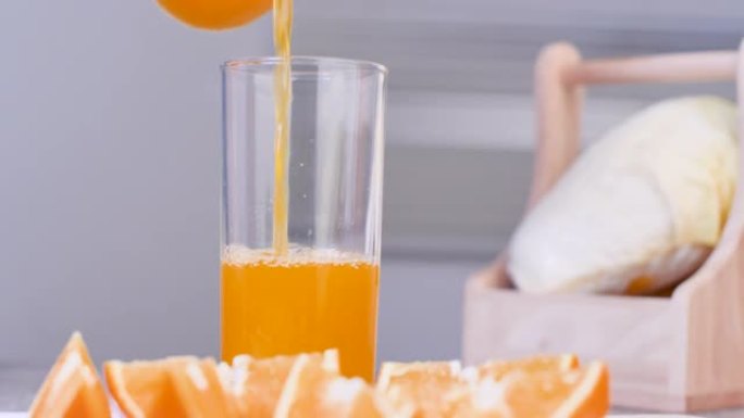 特写镜头将橙汁倒入桌子上的透明玻璃中，橙子切成薄片，放在模糊的前景上。