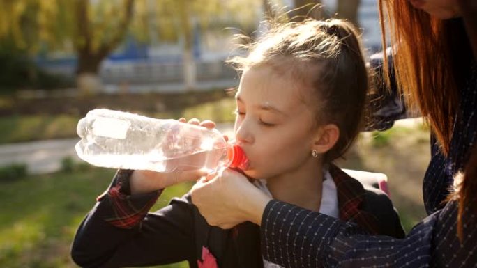 穿着校服的小女孩用塑料瓶喝水。母亲调整女儿的领带，女孩用塑料瓶中的水解渴。