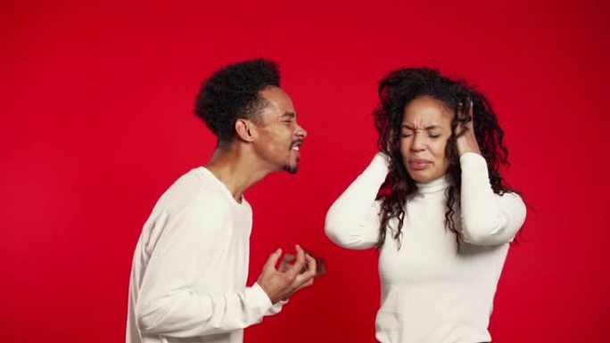 在工作室里，年轻的非洲男子在红色背景上对妻子或女友大喊大叫。无聊的女人用手遮住耳朵。冲突的概念，关系