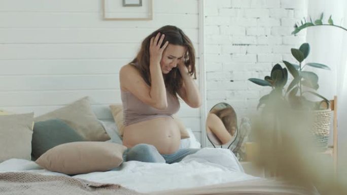 沮丧的孕妇在卧室里哭泣。前卫的肚子妈妈在床上扔枕头
