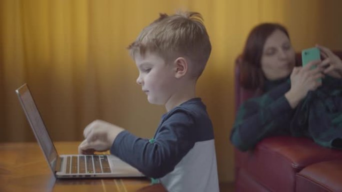 金发碧眼的学龄前男孩在笔记本电脑上打字，微笑着看着电脑屏幕。背景是手机躺在沙发上的母亲。孩子坐在桌旁
