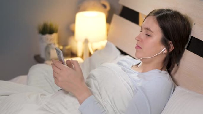 女孩在睡觉前听音乐和使用电话。年轻迷人的女人独自躺在卧室的床上，她戴着耳机听音乐，翻阅手机屏幕。特写