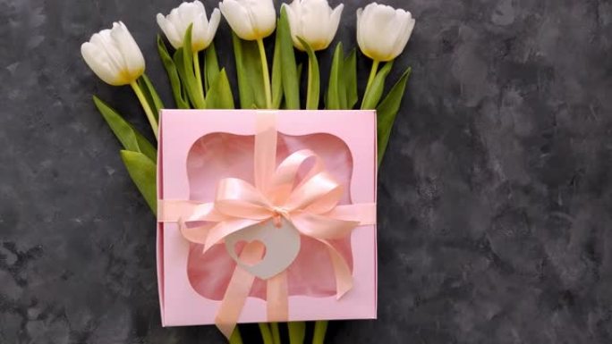 白色郁金香花，带粉色礼品盒和深色背景平板上的心卡标签。4k视频3月8日快乐女性母亲节概念。春天的花束