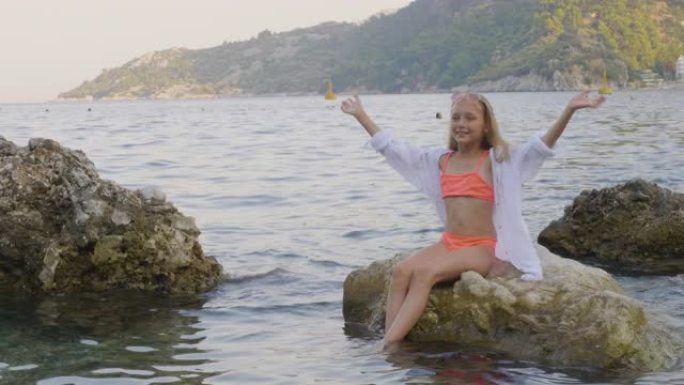 可爱的女孩少年在海石上摇着长发。穿着泳衣和束腰外衣的漂亮女孩在石质海滩的海水中摇着长发拍照。