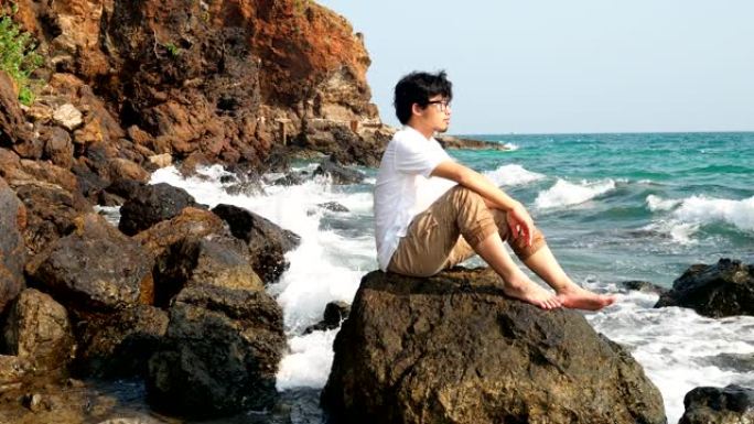 年轻轻松的亚洲男子坐在海边的岩石上