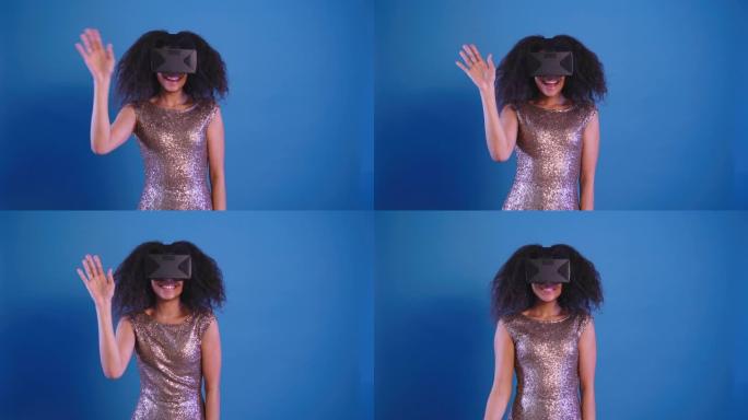 穿着闪亮晚礼服的微笑非洲卷发女人在蓝色背景上孤立的虚拟现实眼镜中迎接某人