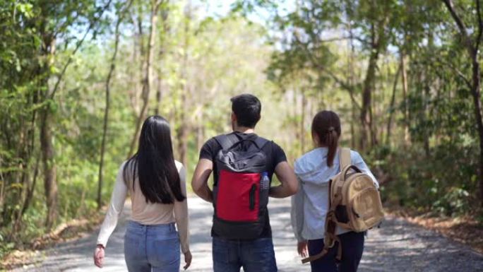 高清慢动作后视组快乐的朋友亚洲女人和男人在夏天在森林土路用背包徒步旅行。放松三个朋友喜欢微笑着散步，