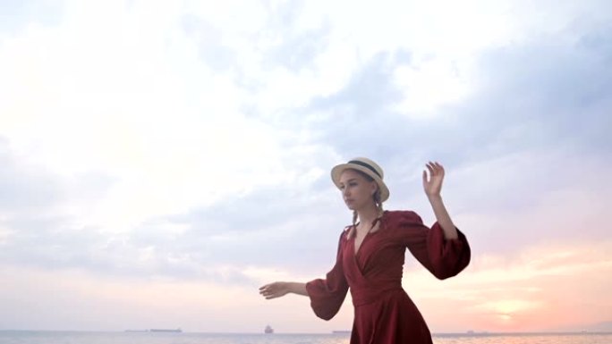 一个迷人的年轻女孩，穿着红色的夏装和草帽，在海浪和日落的背景下，小心翼翼地爬行在岩石海岸的石头上。