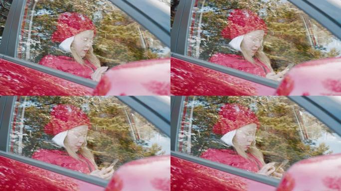 年轻女子在红色车内使用手机。戴着红色帽子和夹克的无忧无虑的女孩在乘客座位上的车内打字智能手机，户外视