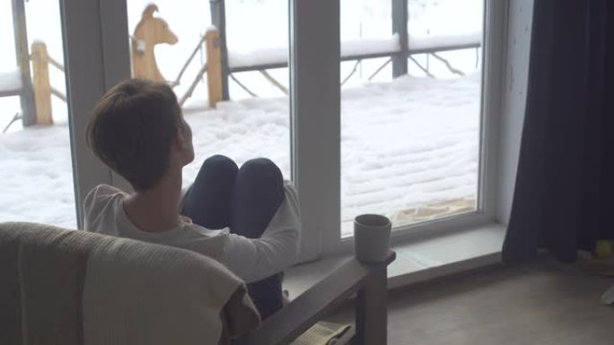 快乐美丽的女孩在一个寒冷的冬日早晨坐在她家窗前的椅子上，看着美丽的景色。一位年轻女子正在喝茶。美丽的