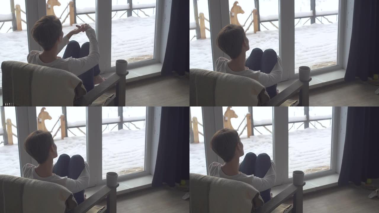 快乐美丽的女孩在一个寒冷的冬日早晨坐在她家窗前的椅子上，看着美丽的景色。一位年轻女子正在喝茶。美丽的