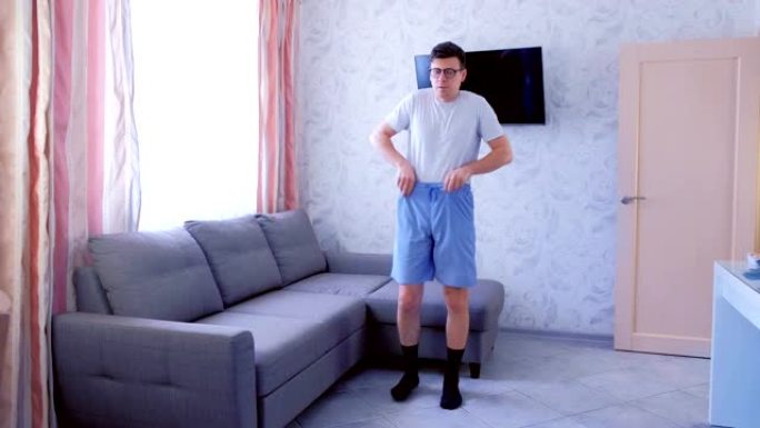 有趣的书呆子男人正在做下蹲和倾斜的腿，用手触摸腿，在客厅在家锻炼。