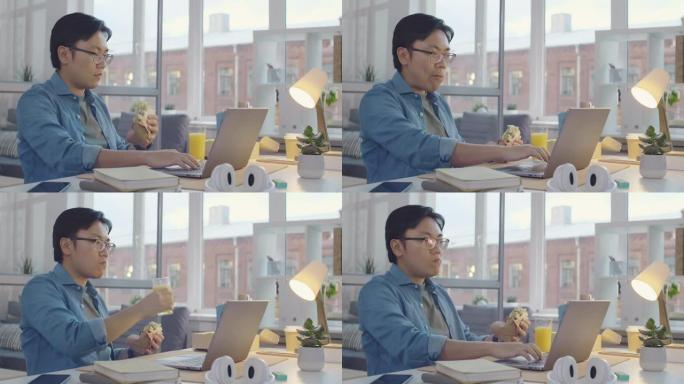 亚洲男子吃三明治并在办公桌前使用笔记本电脑
