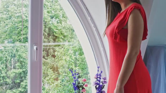 Decade废的年轻女子穿着优雅的红色连衣裙站在窗户旁边，微笑着