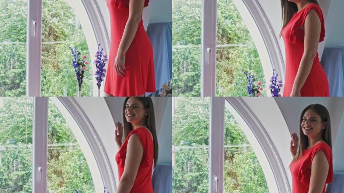 Decade废的年轻女子穿着优雅的红色连衣裙站在窗户旁边，微笑着