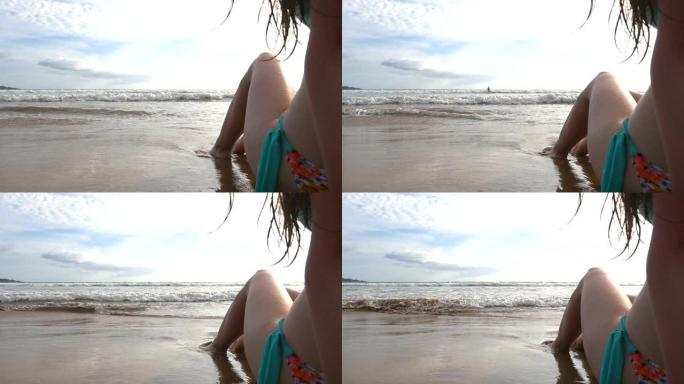 穿着比基尼的晒黑女孩坐在海岸上，在阳光明媚的日子晒日光浴。海浪在海岸上冲刷着年轻的女性脚。背景风景优